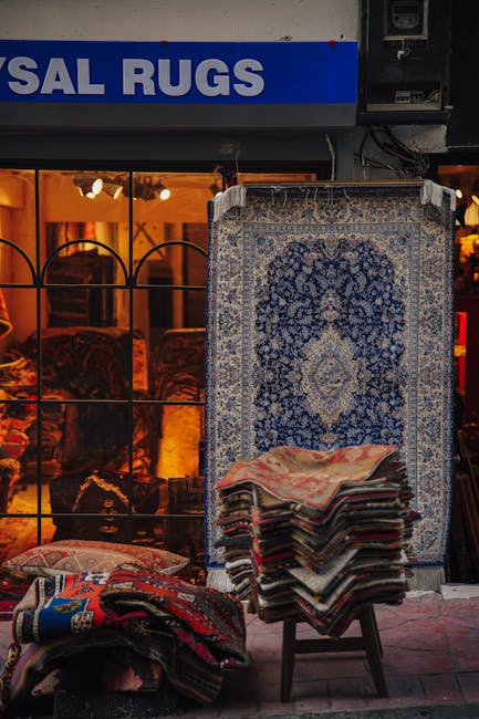A person wearing a blue and white striped shirt is kneeling on a wooden floor, performing surface cleaning or deep cleaning on a patterned red, blue, and cream area rug. The person's hands are placed 