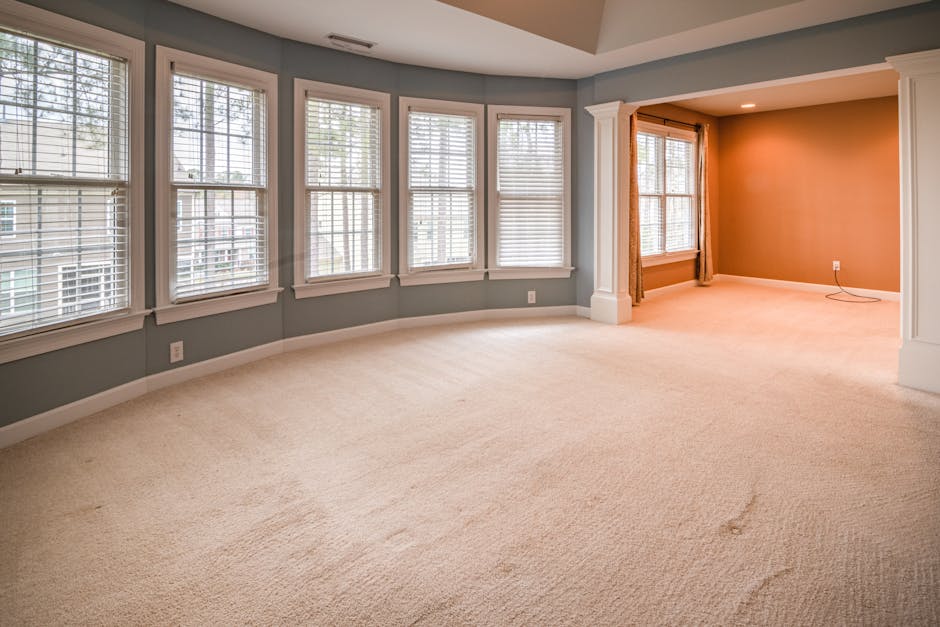 A spacious, empty residential living room featuring a cream-colored carpet that appears clean and well-maintained. The room has large white-framed windows fitted with horizontal blinds, allowing natur