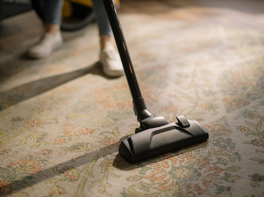 Close-up of a modern vacuum cleaner head being used on a patterned oriental-style rug in a well-lit room, with a person's white sneakers and dark trousers visible in the background. The surface appears clean with a slight sheen, indicating a recent deep cleaning or sanitisation process. The vacuum cleaner is black with a slim nozzle, positioned centrally on the rug, which features intricate floral motifs in muted tones of beige, green, and orange. The scene emphasizes surface cleaning and maintenance within a domestic setting, consistent with Harrow Carpet Cleaners' services for carpet and upholstery cleaning in South Harrow HA2. The room’s bright lighting highlights the detailed pattern and cleanliness of the flooring.