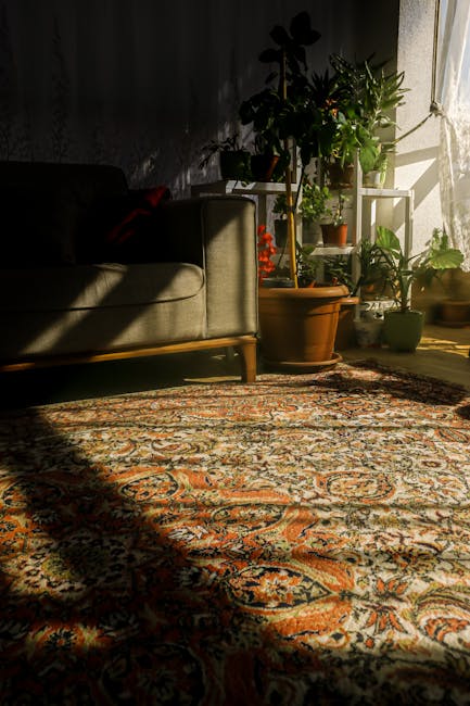A close-up view of a patterned carpet with intricate floral and geometric designs in shades of orange, beige, and blue, situated in a well-lit living room corner. The carpet appears clean and plush, reflecting natural sunlight streaming in from a nearby window. To the left, part of a green upholstered sofa with wooden legs is visible, partially shaded. On the right, several potted plants of various sizes and types are placed on a white, multi-tiered plant stand, some with green foliage, illuminated by the sunlight, creating bright highlights on their leaves. The shadows cast on the wall behind and on the carpet suggest a warm, inviting atmosphere. This setting is typical for a domestic environment where routine surface cleaning and maintenance contribute to hygiene and comfort, aligning with Harrow Carpet Cleaners' expertise in deep cleaning and sanitation for homes. The image emphasizes clean, well-maintained surfaces and the use of natural light to enhance the aesthetic appeal of the space.
